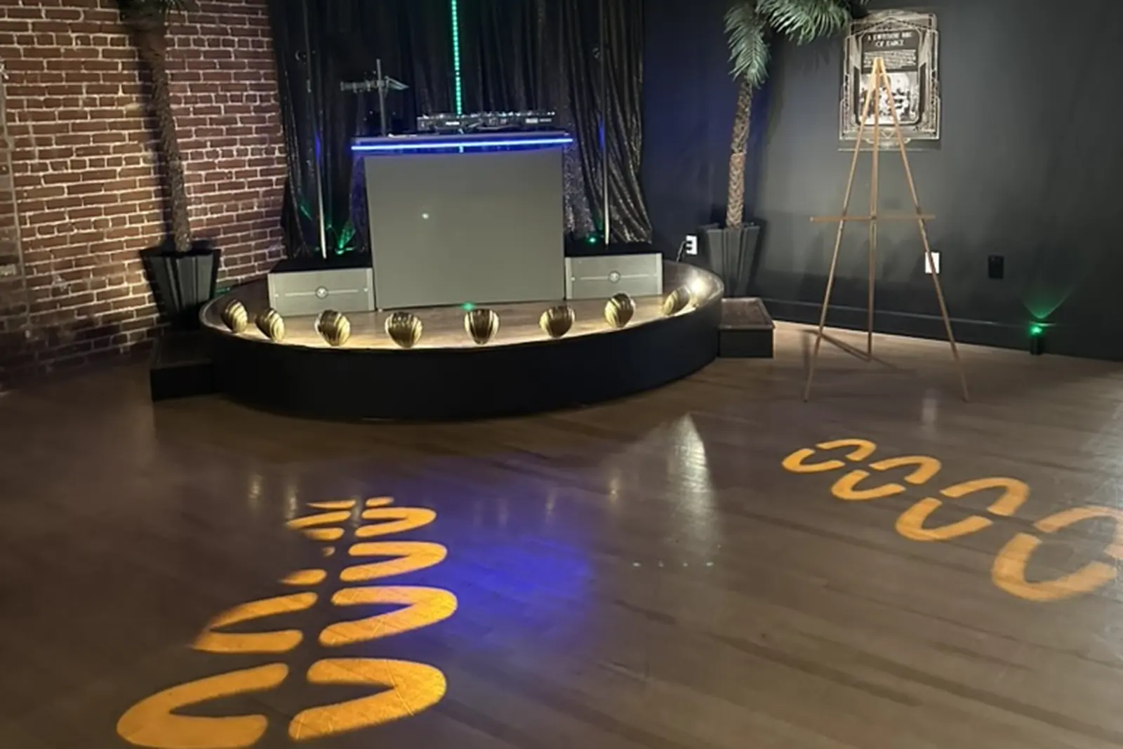 A DJ booth on a low round stage in a brick-walled event space, lit green from behind, with gold patterned lighting projected across the wood floor and an easel sign beside the stage.