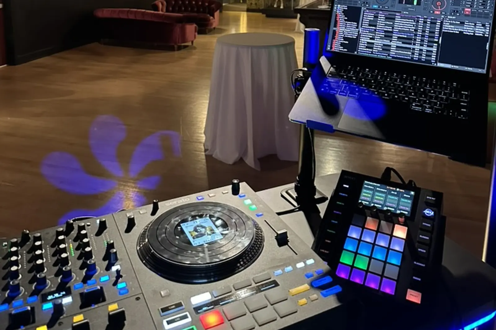 The view from behind a DJ controller across an event room: chandeliers, a velvet sofa, a draped cocktail table, and a purple pattern thrown across the floor.