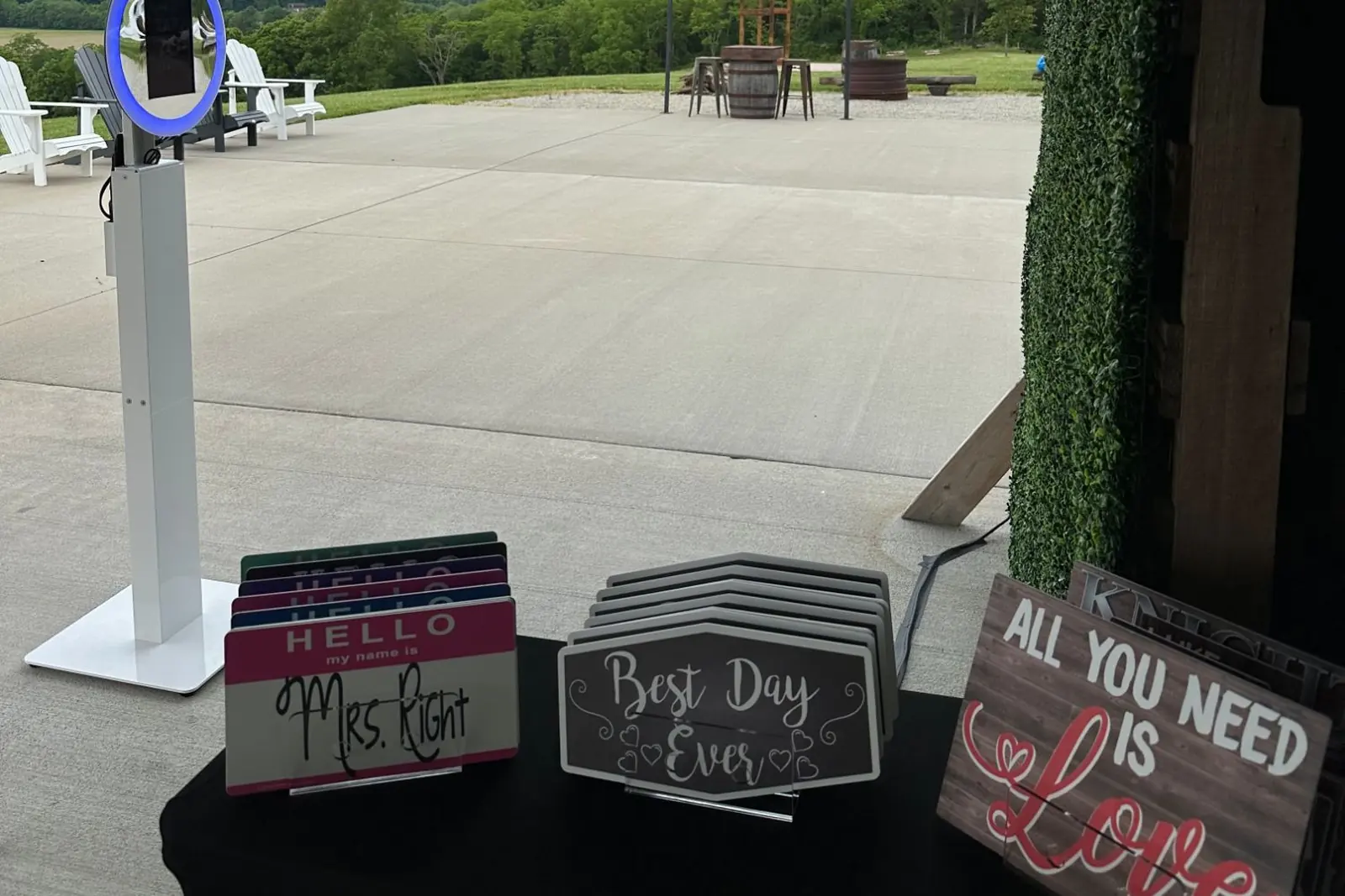 An open-air photo booth on a slim white stand with a blue ring light, set up in a barn doorway looking out over a patio and wooded hills, with a table of props in front: name badges, chalkboard signs and an All You Need Is Love board.