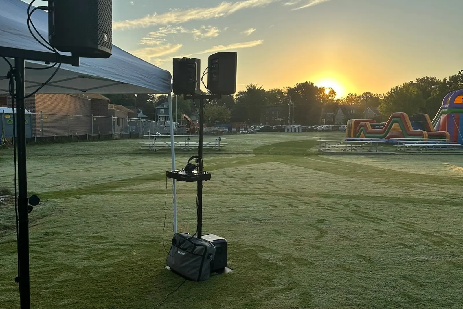 Two loudspeakers on stands beside a pop-up tent on an open sports field at sunrise, powered by a battery pack sitting on the grass, with inflatables and bleachers in the distance.