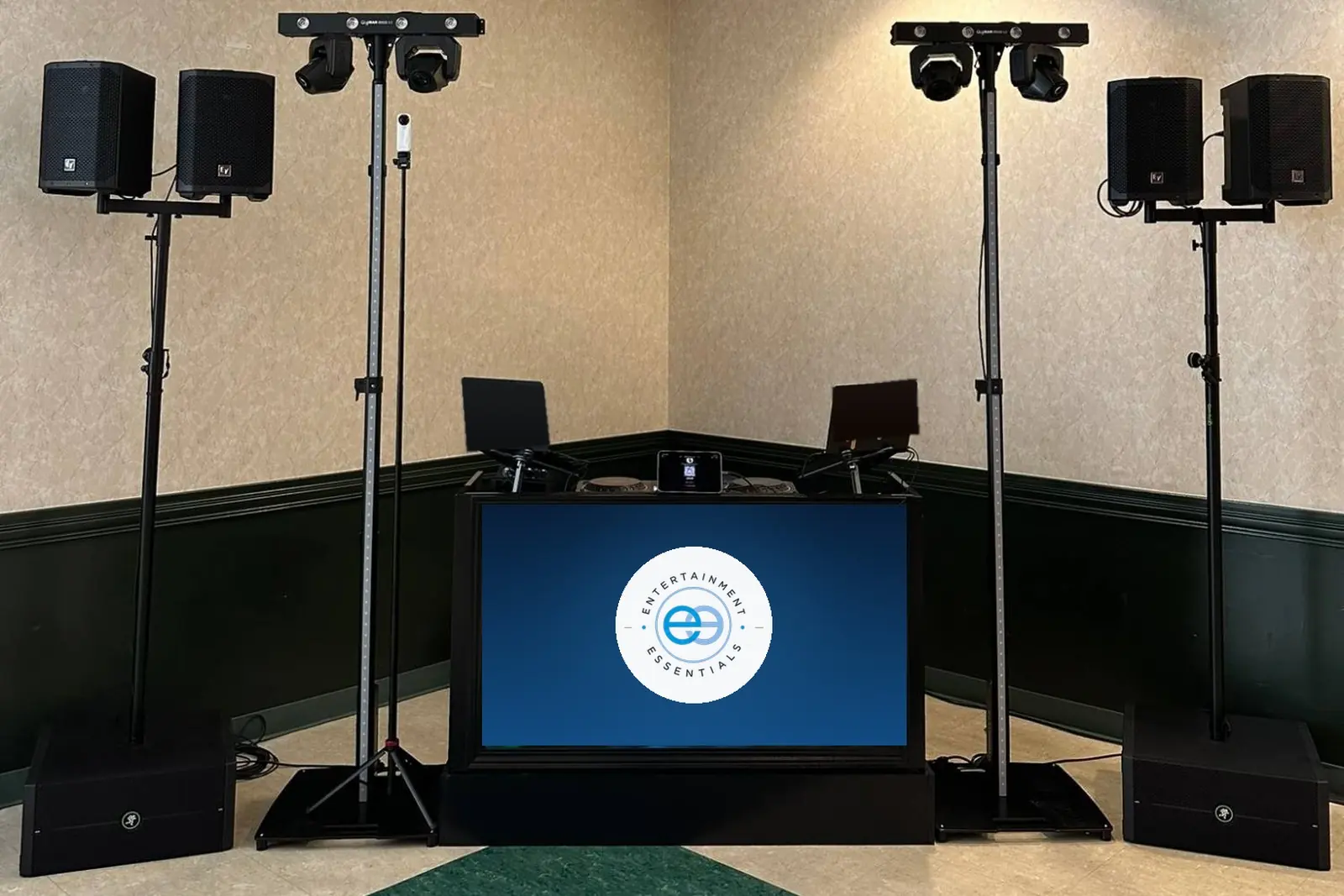 A school dance setup in a corner of a hall: two pairs of speakers on stands, moving-head lights on lighting towers, subwoofers on the floor, and a DJ booth with a lit screen carrying the Entertainment Essentials mark.