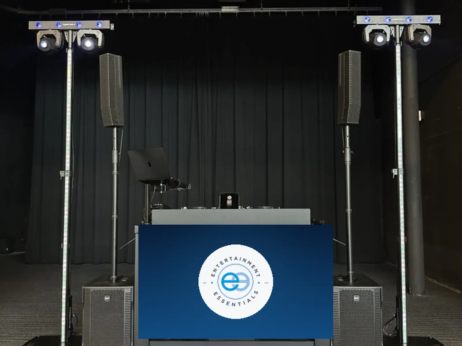 A wedding reception setup against black drape: two line-array speakers on stands, moving-head lights on LED towers either side, and a DJ booth with a large screen showing the Entertainment Essentials mark.